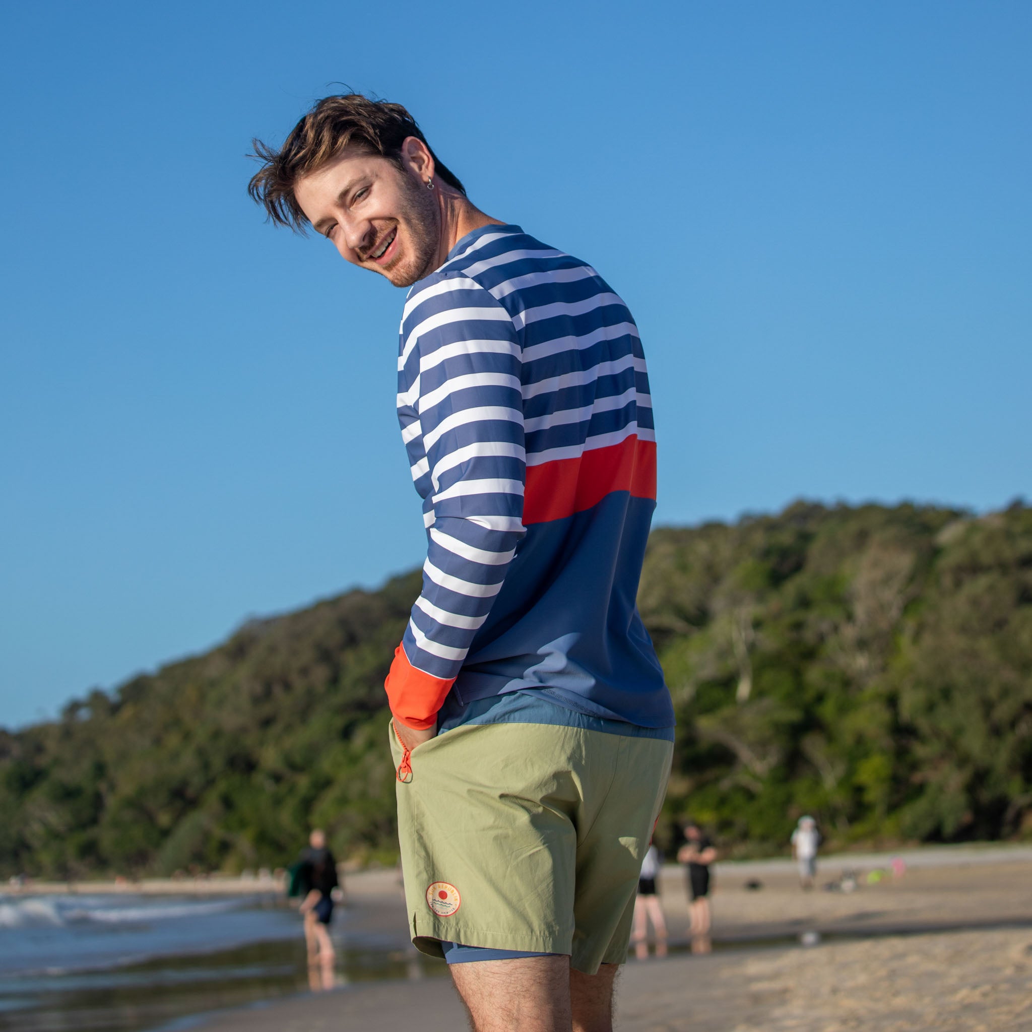 Man standing on sand smiling, wearing mens beach wear swim shirt with long sleeves.