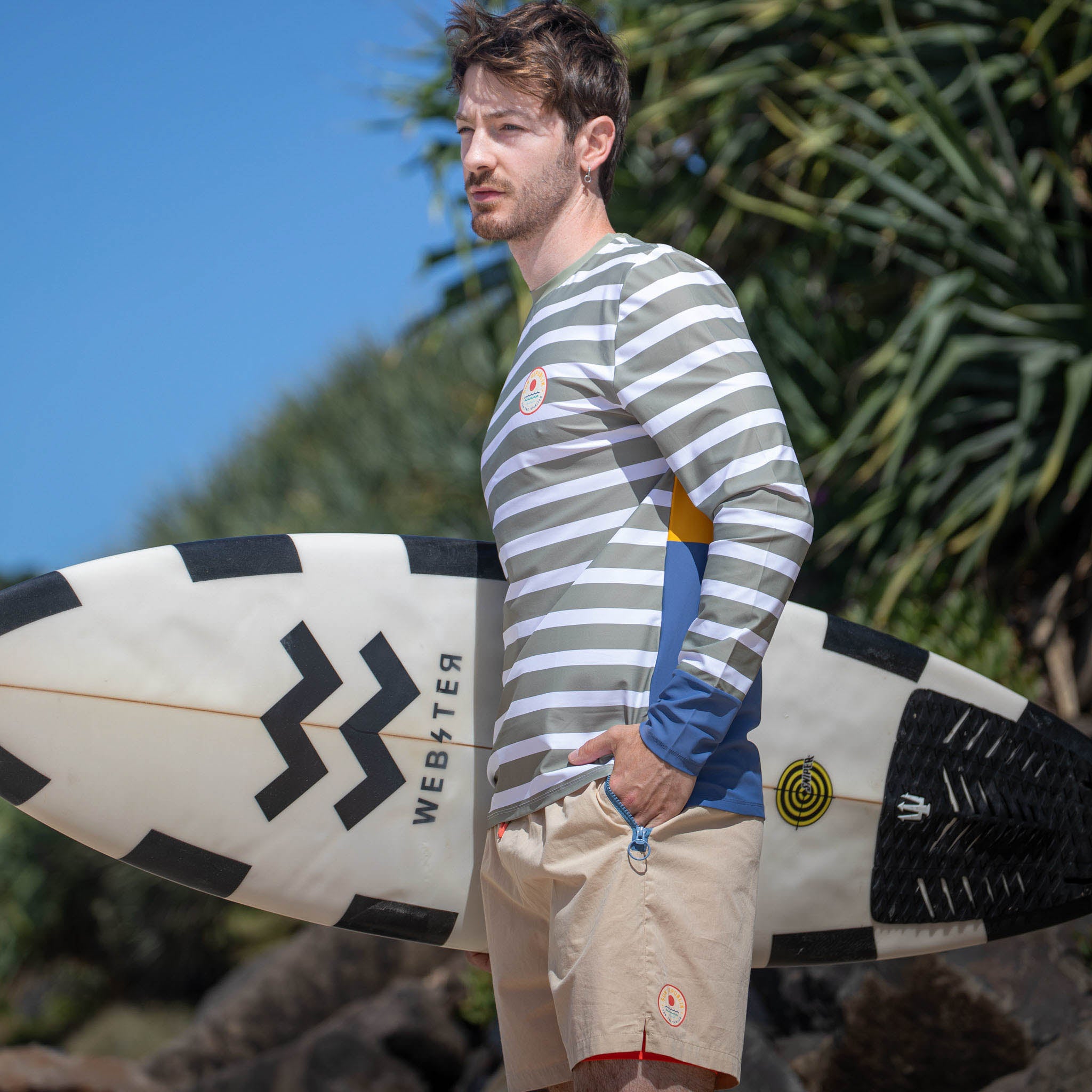 Man on beach wearing men swimwear swim shirt holding surfboard.