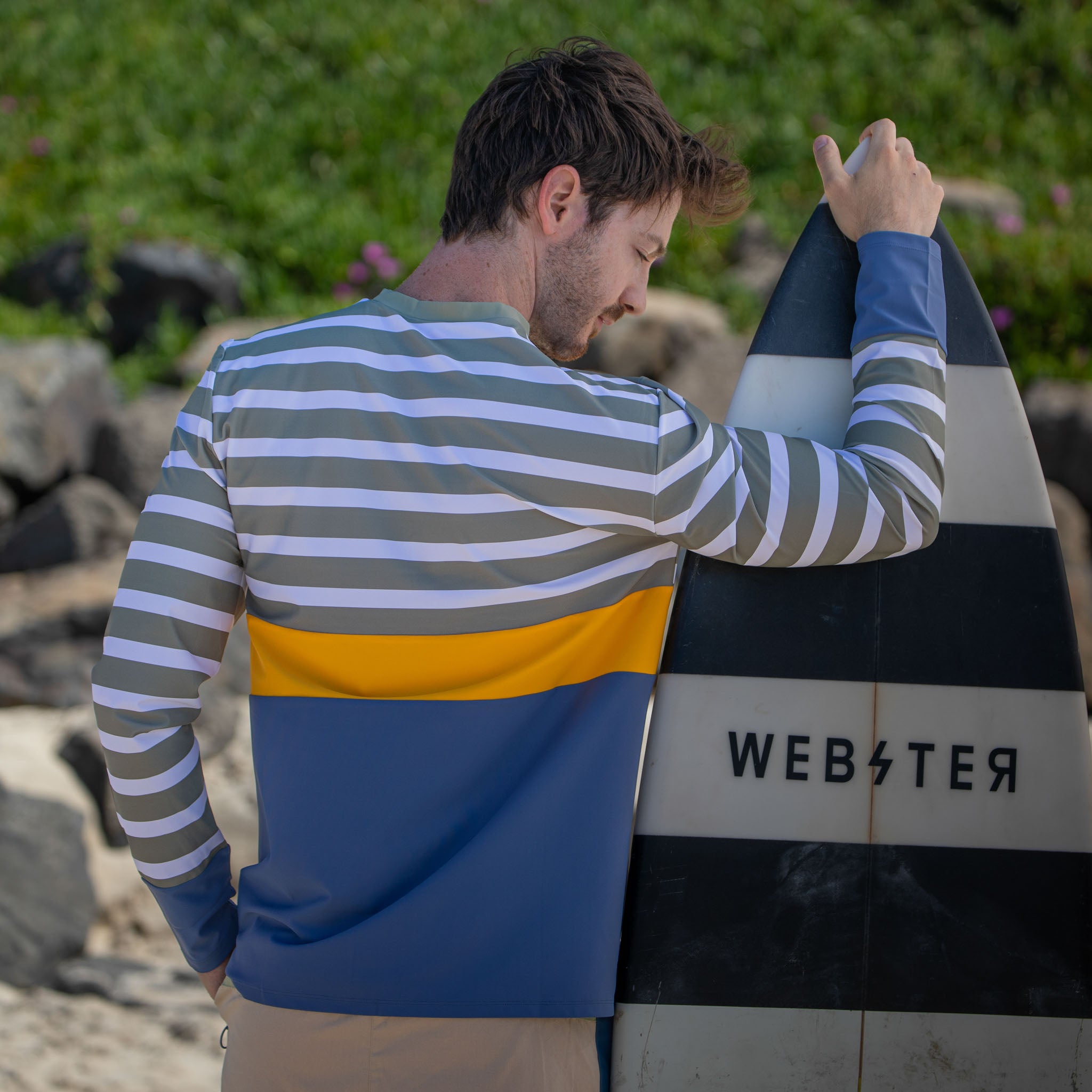 Back view of man with surfboard wearing men swimwear rash top, long sleeve.