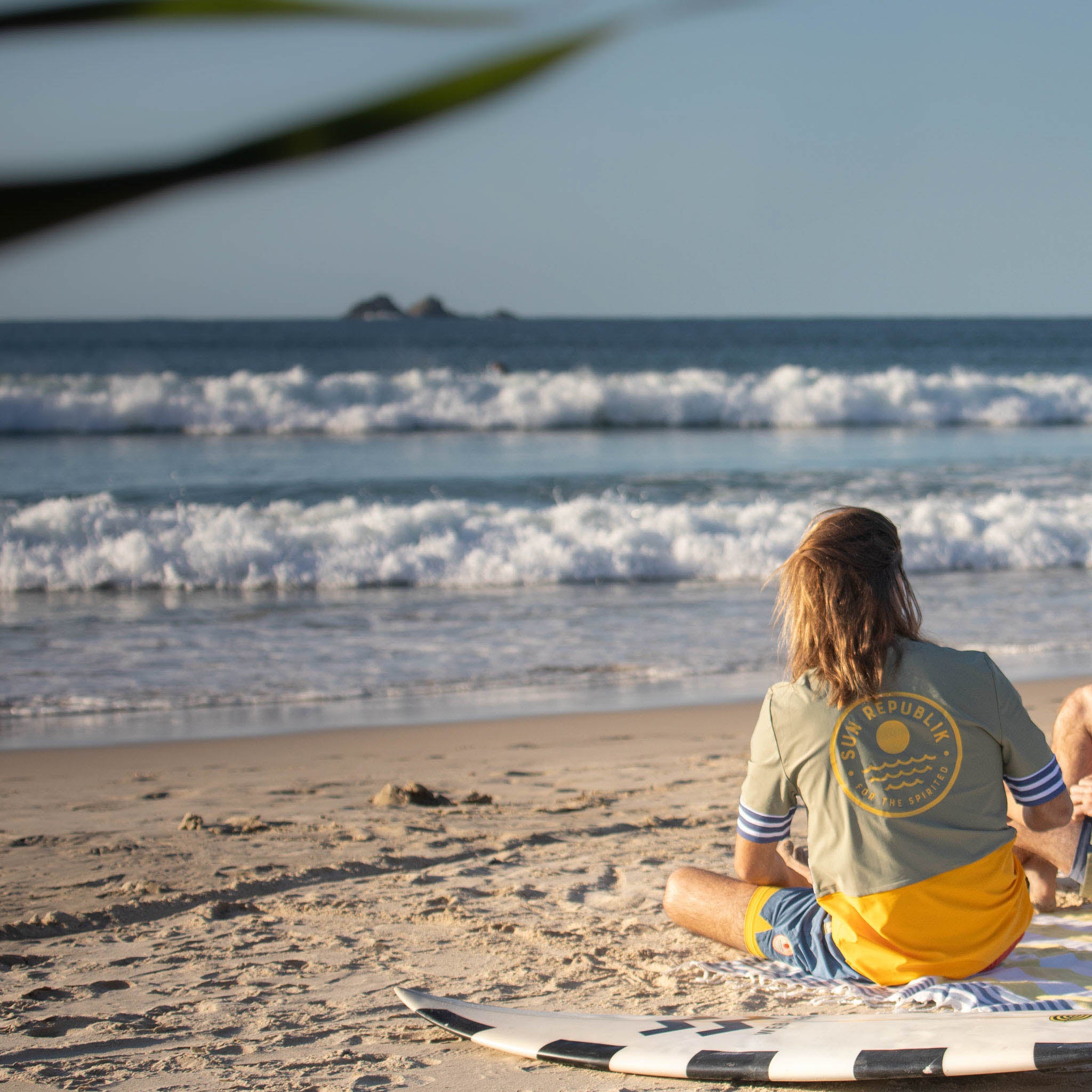 Man sitting on beach wearing men swimwear, rash guard top. Short sleeve and back logo.