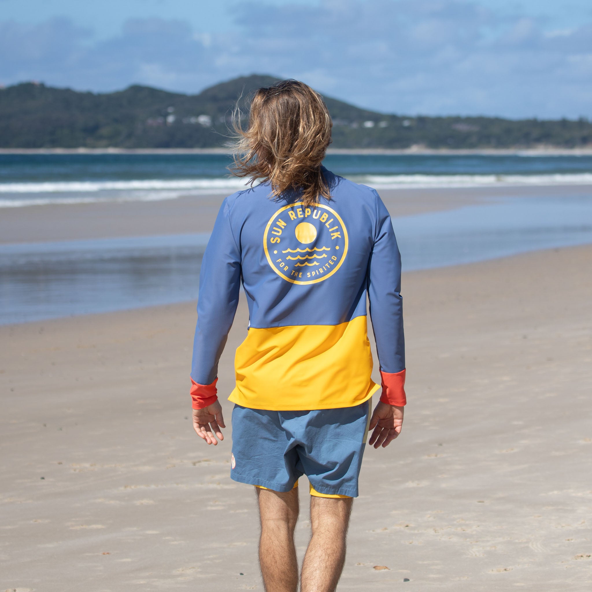 Back view of man walking wearing mens beach wear swim top with long sleeve and logo.