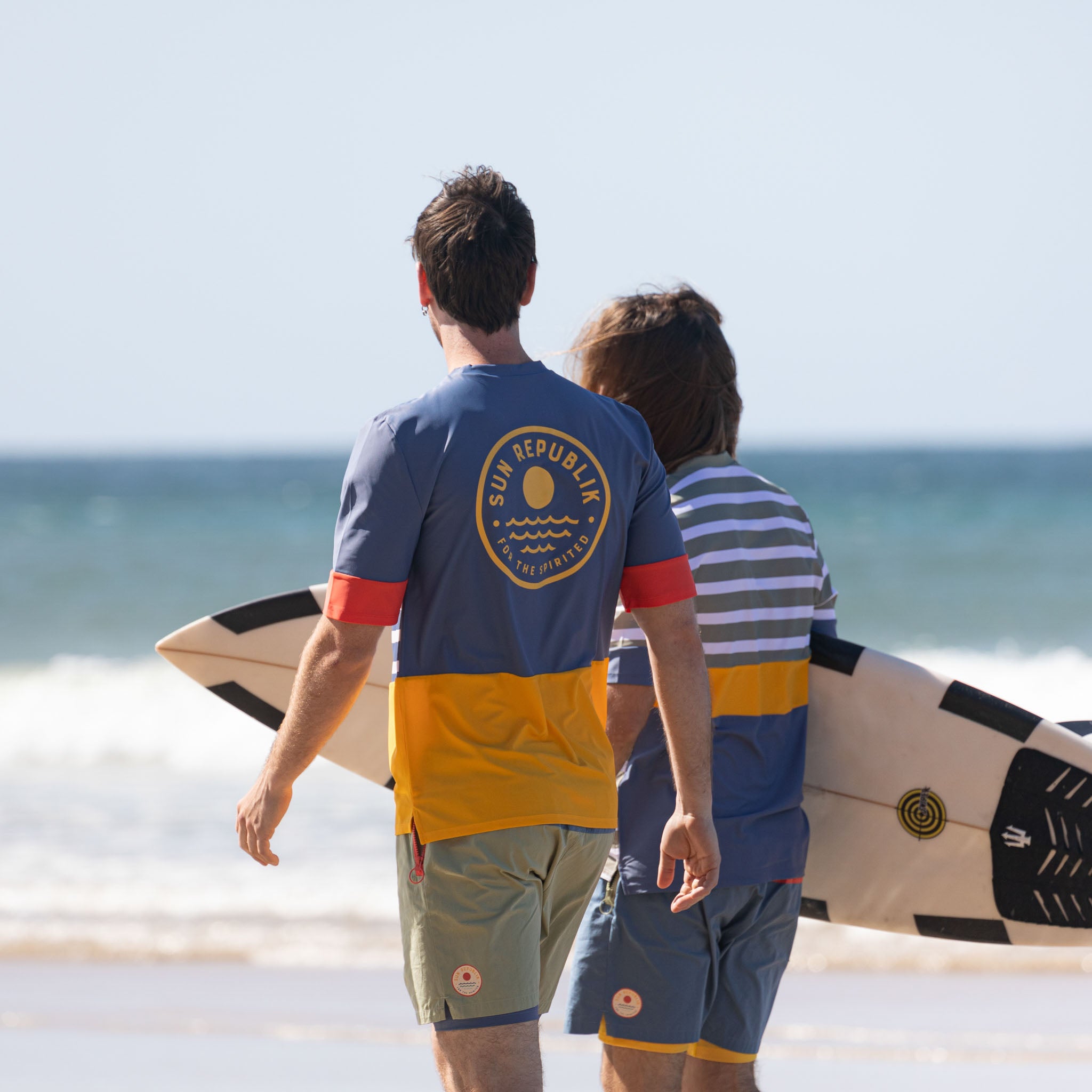 Men walking on beach wearing men swimwear rashie top and shorts. Sun republik logo on back.