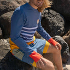 Man sat on rocks wearing men swimwear, long sleeve swim shirt and beach shorts.